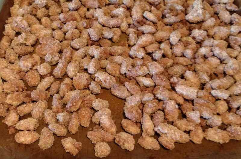 Candied almonds spread out on a parchment-lined tray, coated in a golden-brown crystallized sugar glaze, cooling and ready to enjoy.