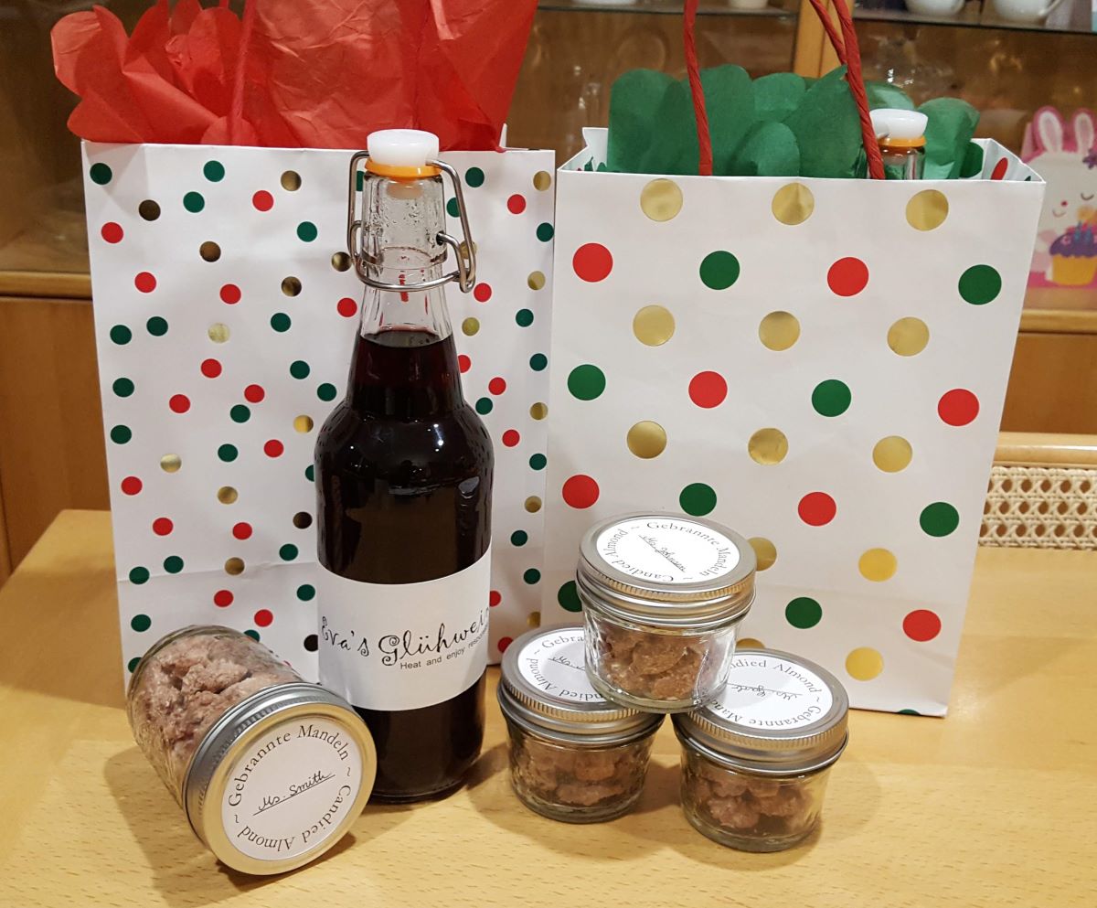 Homemade holiday gift set featuring a bottle of Glühwein and several jars of candied almonds, displayed in front of festive gift bags with red, green, and gold polka dots.